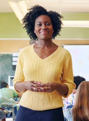 A smiling teacher in a yellow sweater stands in a classroom