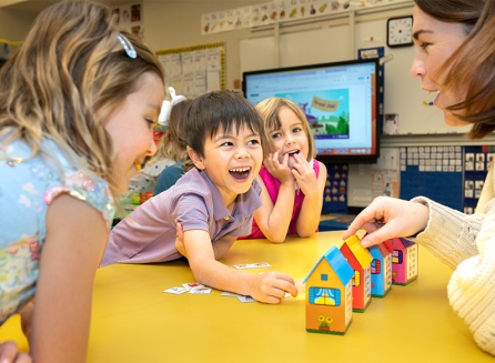 lower school students smiling while interacting with teacher who has a variety of toy houses
