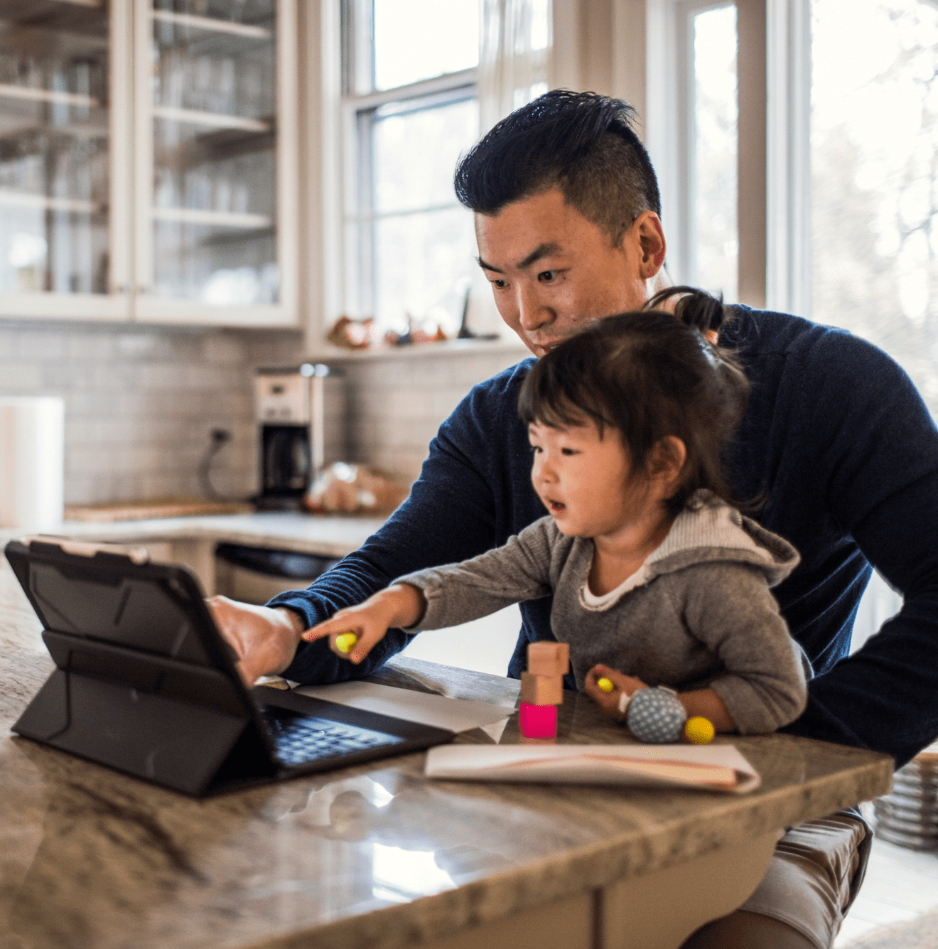 Man sits at table with young daughter in lap both pointing at tablet screen and smiling.