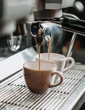 Fresh espresso streaming from a professional coffee machine into two white ceramic cups on a metal drip tray.
