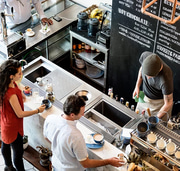 Overhead view of customers at a marble cafe counter being served by a barista, with a chalkboard menu in the background.