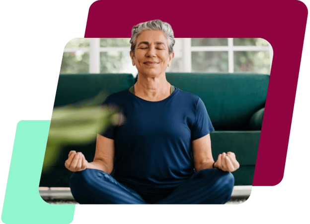 Uma imagem de uma mulher branca de cabelo curto grisalho, meditando de olhos fechados sentada no chão de uma sala, de blusa azul e calça legging também azul.