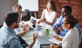 Group of male and female colleagues sit around meeting table having a conversation.