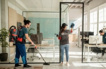 Image of 3 people cleaning an office, one hoovering the floor, one cleaning a window partition and someone cleaning a desk