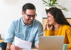 man and woman sitting at desk. man is holding paper and looking at computer. woman is looking at man
