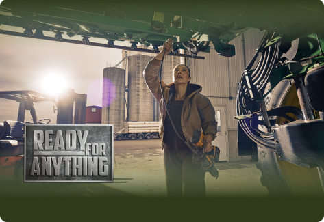 A woman in work overalls repairs a large agricultural machine in a farm workshop, with silos and a forklift in the background 