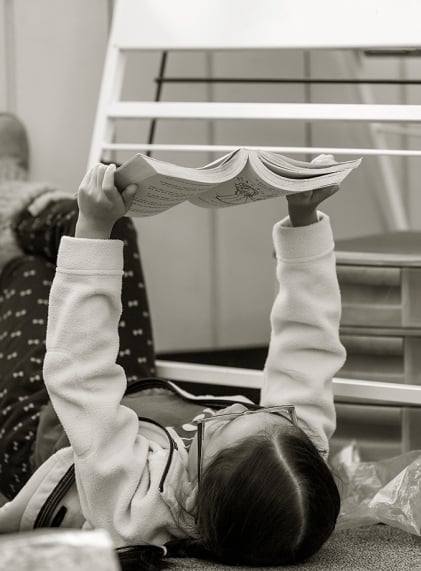 Elementary age girl in a classroom lays on her back, reading a book she holds at arm's length into the air