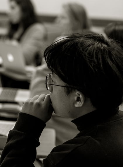 A student sits in the foreground with their hand on their chin, listening intently to the professor, open laptop in front of them. Other students can be seen in the background, blurred.