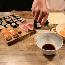 Sushi on a cutting board with a person pouring sauce in a bowl 