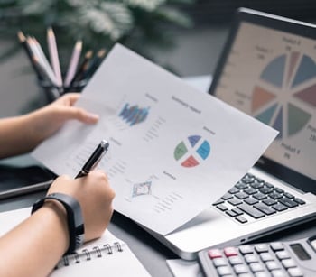 Hand holding paper with graphs and a graph on the screen of a laptop with a hand writing notes