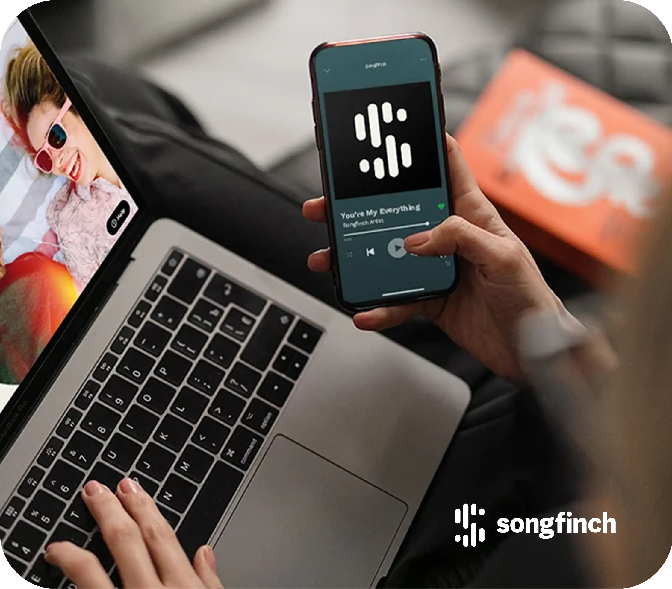 Hand holding a phone with a music app open next to a laptop keyboard, Songfinch logo in the corner.