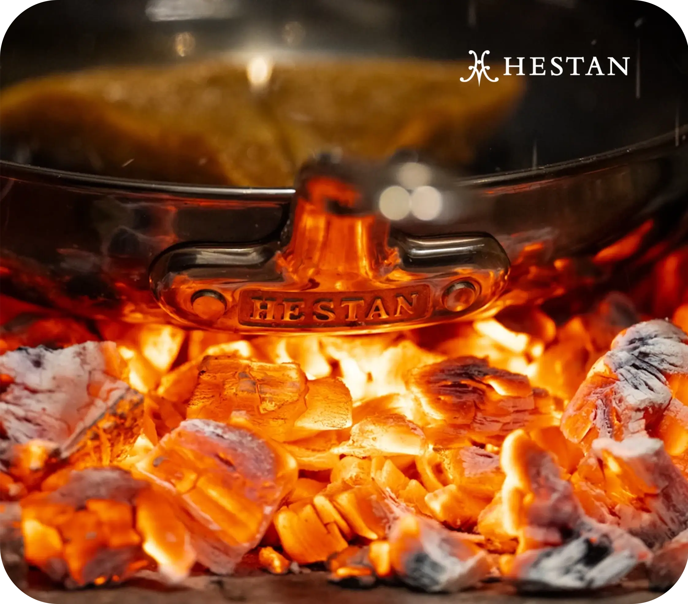 Close-up of a Hestan pan over glowing coals, Hestan logo visible.