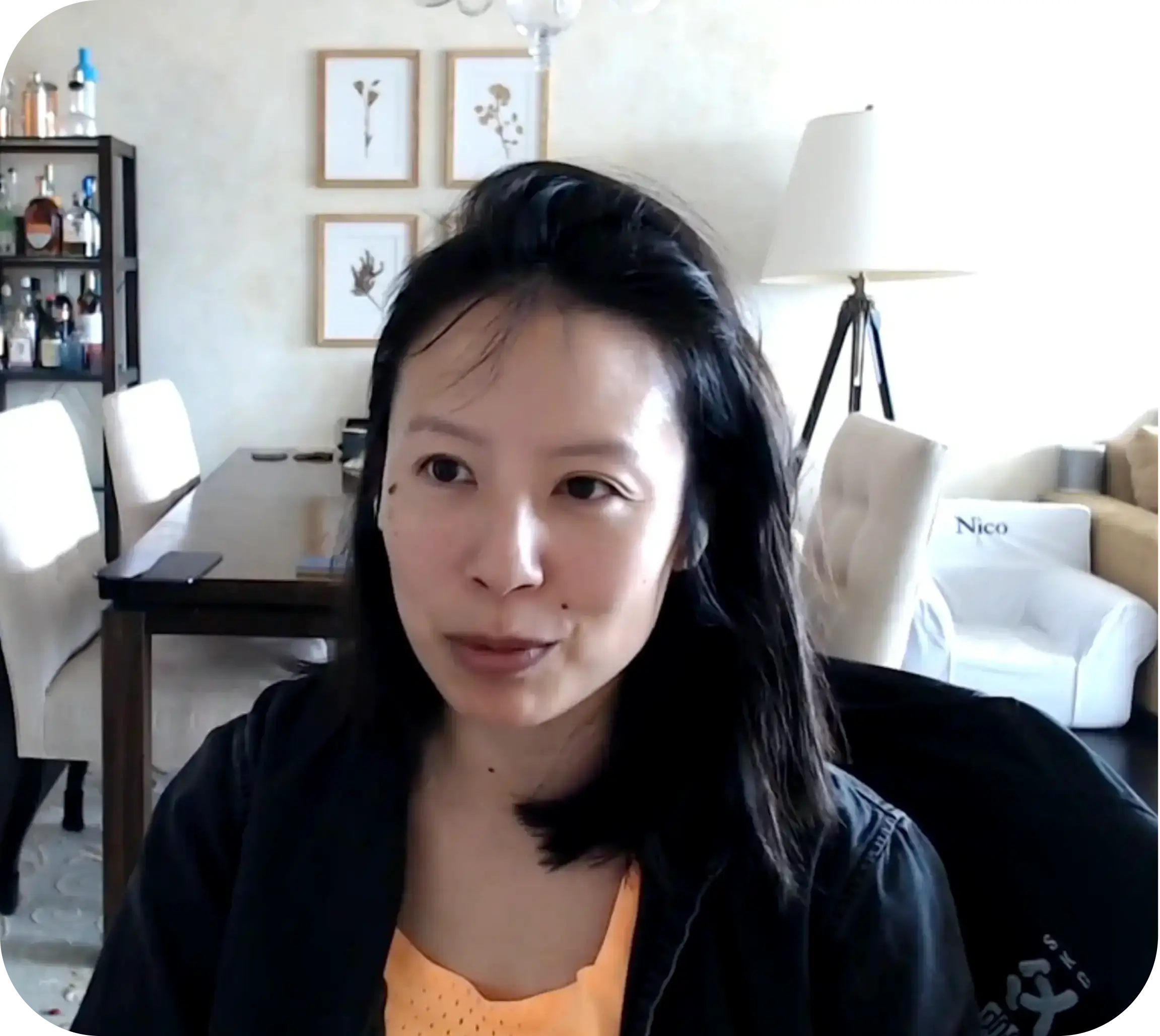 Woman speaking on a webcam in a bright living room with dining table and shelving behind her.