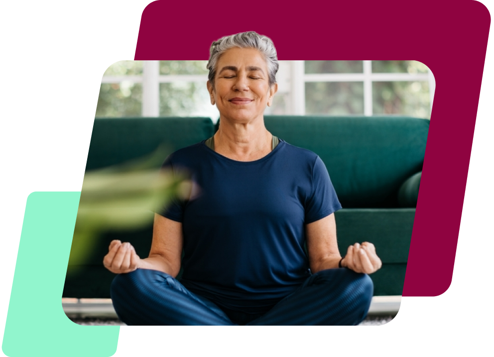 Uma imagem de uma mulher branca de cabelo curto grisalho, meditando de olhos fechados sentada no chão de uma sala, de blusa azul e calça legging também azul.