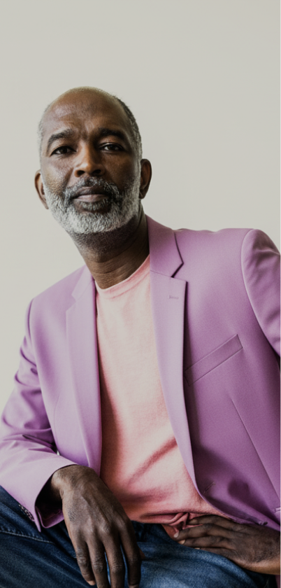 man sitting. he is wearing a lavender  blazer and jeans. he has a gray beard
