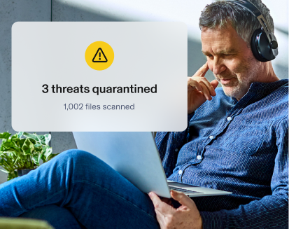Man using headphones and laptop. Aura app overlay shows 3 threats quarantined with 1,002 files scanned.