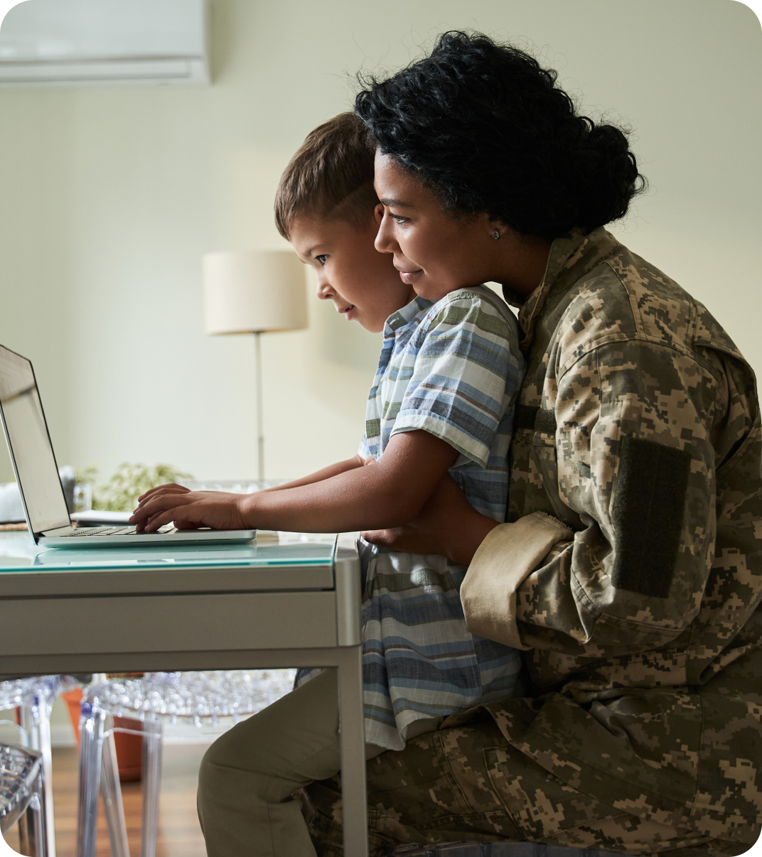 A woman in military uniform sits with a young child on their lap, both looking at a laptop together at a home desk.
